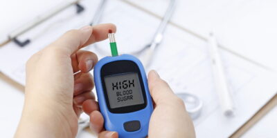 Hand holding a blood glucose meter measuring blood sugar, the background is a stethoscope and chart file